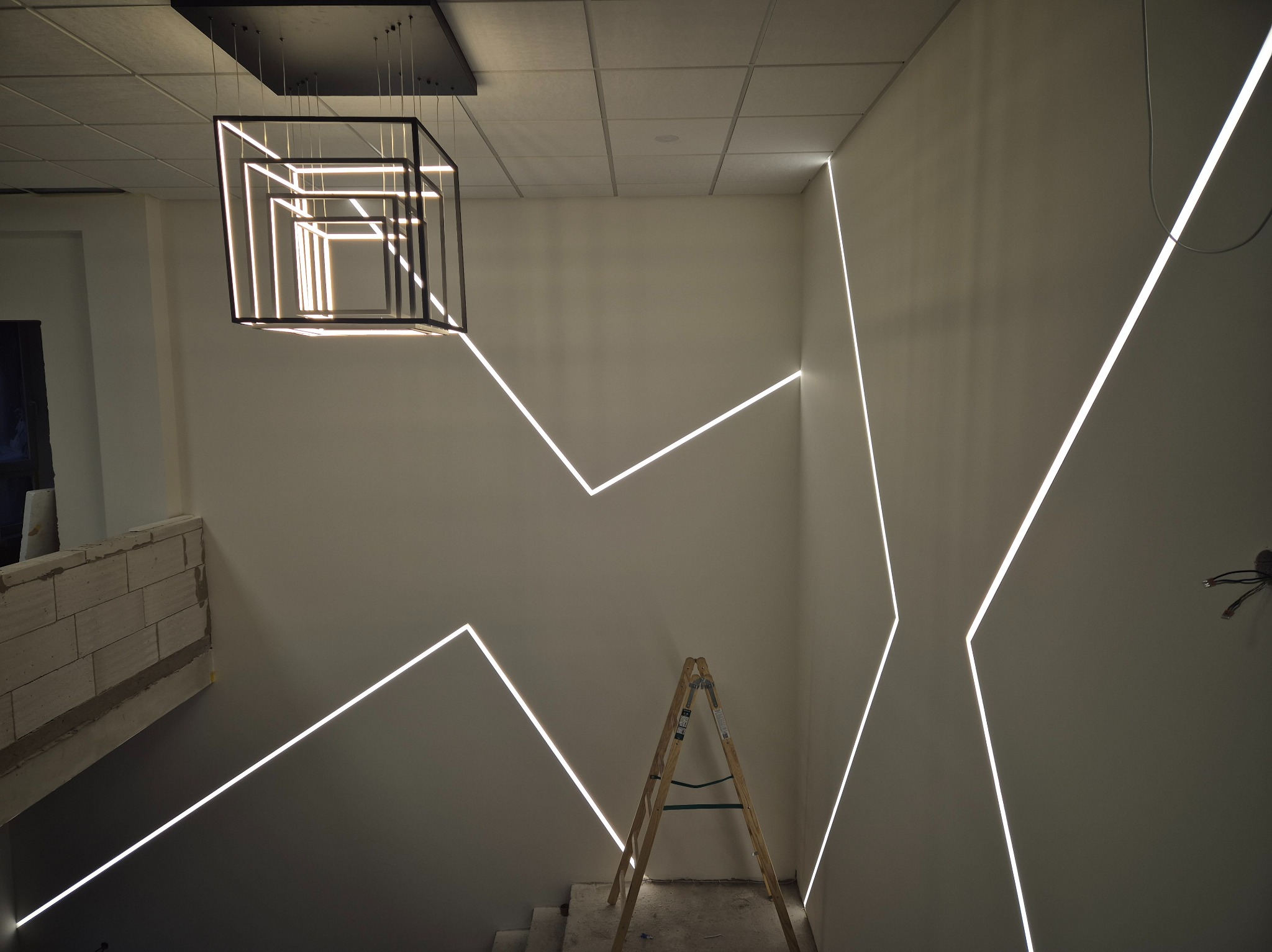 Nowoczesne oświetlenie schodów z geometrycznymi liniami LED na ścianach i designerską lampą wiszącą w kształcie sześcianu. Widoczna drabina malarska.