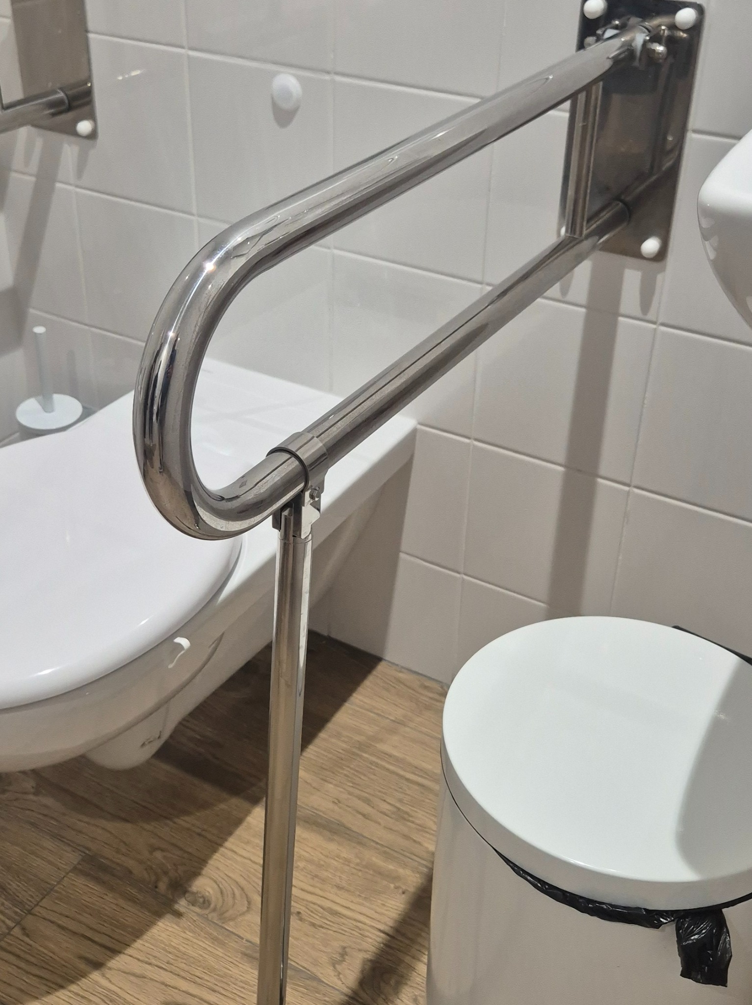 Zamontowana, chromowana poręcz dla niepełnosprawnych w łazience z białymi płytkami i drewnopodobną podłogą. Widoczny fragment toalety i kosza na śmieci.