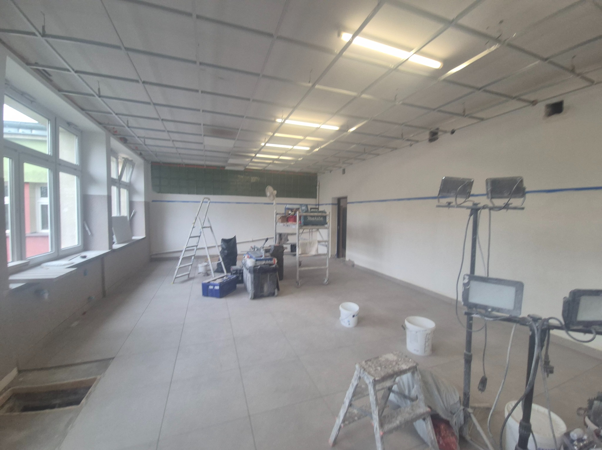 Wnętrze w trakcie prac wykończeniowych: widoczne rusztowania, drabiny, narzędzia malarskie, oświetlenie i przygotowane ściany do malowania. Częściowo położona podłoga.