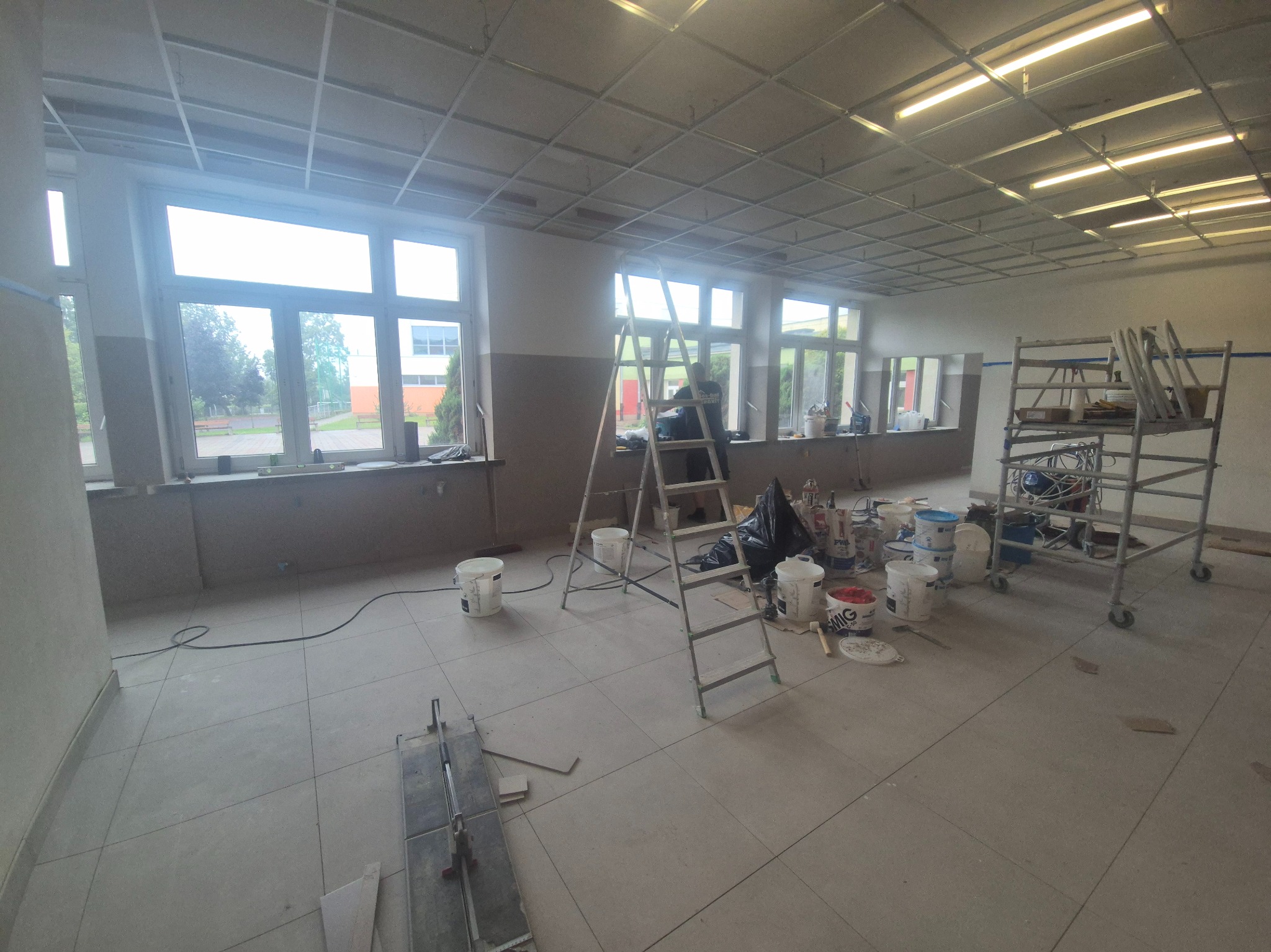 Wnętrze w trakcie prac wykończeniowych: drabina, rusztowanie, wiadra z farbą, narzędzia glazurnicze na podłodze z płytek. Widoczne okna i częściowo zamontowany sufit podwieszany.