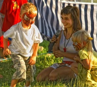 Animator dla dzieci z mikrofonem podczas malowania twarzy dzieciom w plenerze. Chłopiec z twarzą Spidermana, dziewczynka z motywem tygrysa. W tle namiot w biało-granatowe pasy.