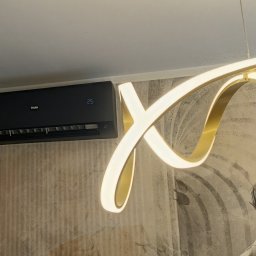 ENEUS Rafał Minierski - Czarny klimatyzator ścienny Haier z cyfrowym wyświetlaczem 25, zamontowany na teksturowanej ścianie z muralem, obok nowoczesnej, podświetlanej lampy.