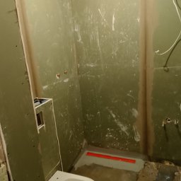 Błażej Ptaszyński - Łazienka w trakcie remontu z zielonymi płytami gipsowo-kartonowymi, częściowo zamontowaną miską WC, liniowym odpływem prysznicowym i widocznymi instalacjami wodno-kanalizacyjnymi.