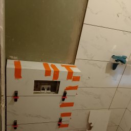 Błażej Ptaszyński - Układanie białych płytek ściennych z marmurowym wzorem w łazience. Widoczna zabudowa stelaża podtynkowego zabezpieczona pomarańczową taśmą i klipsami poziomującymi.