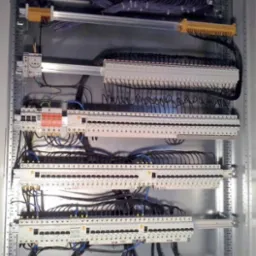 Rozbudowana szafa sterownicza z licznymi modułami, listwami zaciskowymi i przewodami w różnych kolorach, zamontowana na szynach w metalowej obudowie.