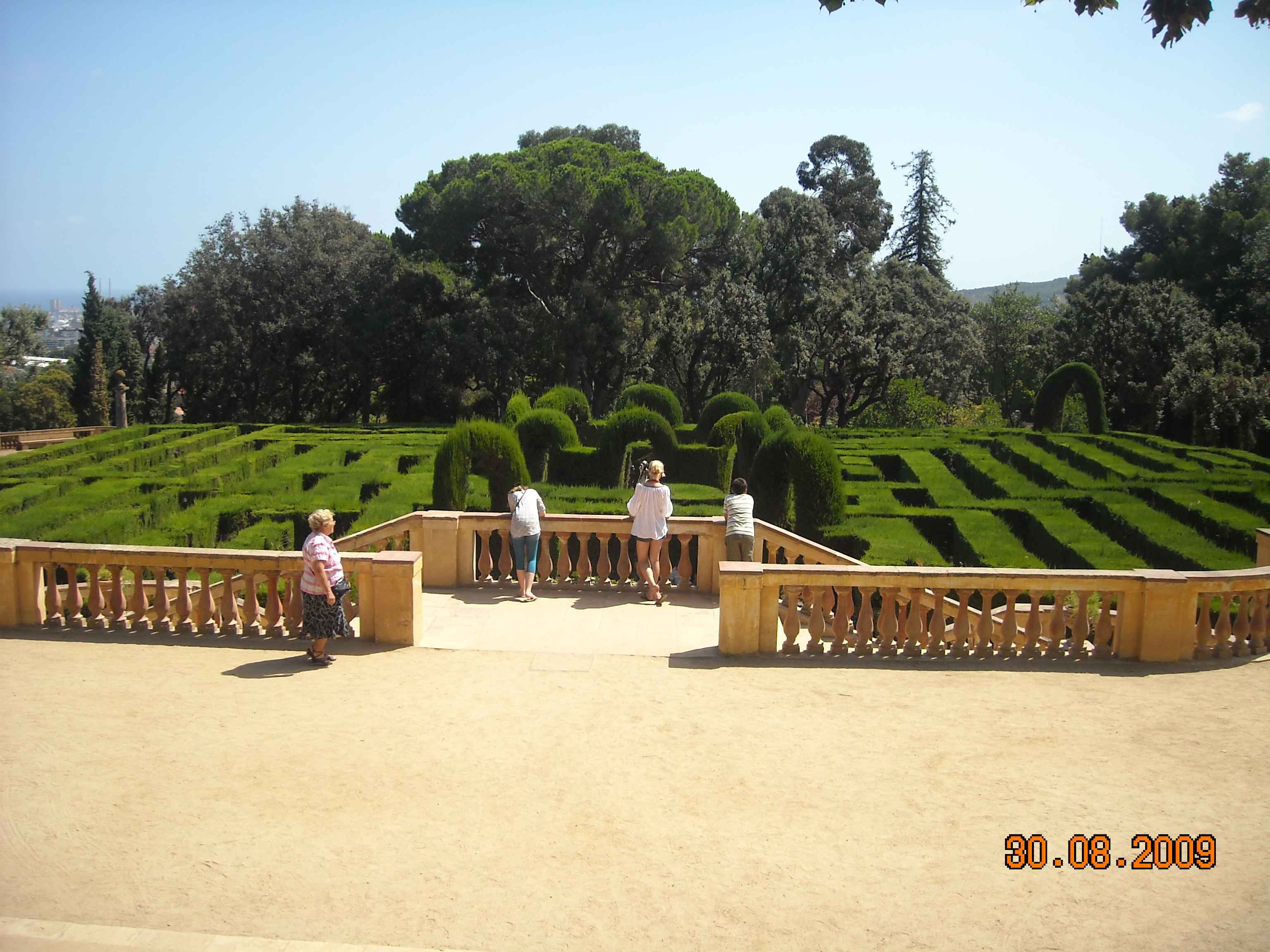 Labirynt z żywopłotu w parku, grupa turystów obserwuje z tarasu, słoneczny dzień.