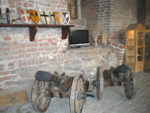 Wnętrze z elementami średniowiecznego wystroju: dwie drewniane repliki armat na kółkach, tarcze herbowe na półce, ceglana ściana i telewizor.