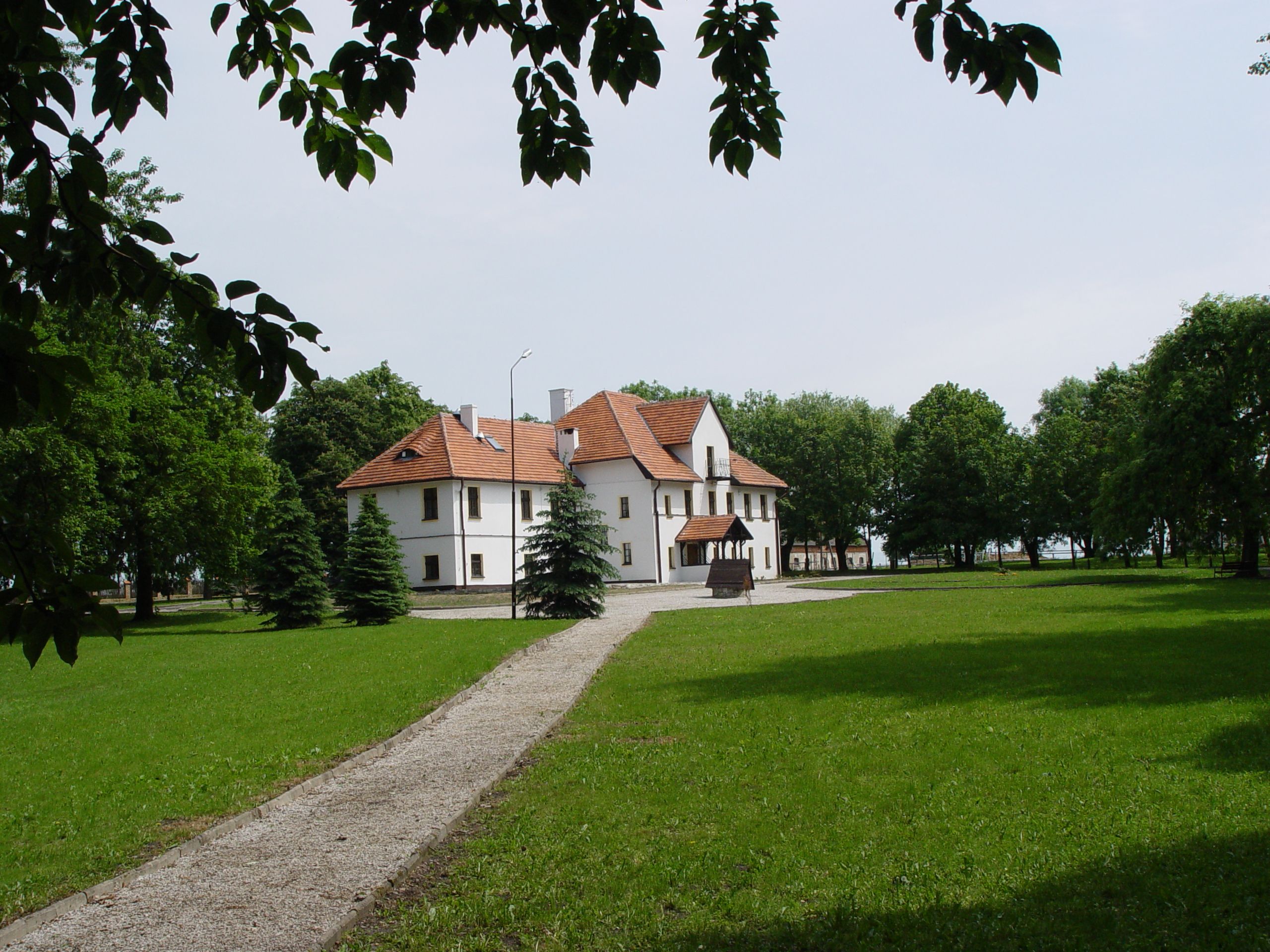 Dwór w stylu klasycystycznym z czerwoną dachówką, otoczony zielonym trawnikiem i drzewami, widok z perspektywy ścieżki prowadzącej do budynku, z liśćmi drzew w górnej części kadru.