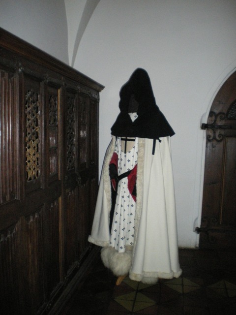 Strój historyczny składający się z białego płaszcza z futrzanym wykończeniem, czarnego kaptura i wzorzystej sukienki, prezentowany na manekinie w zabytkowym wnętrzu z ciemnymi, drewnianymi panelami...