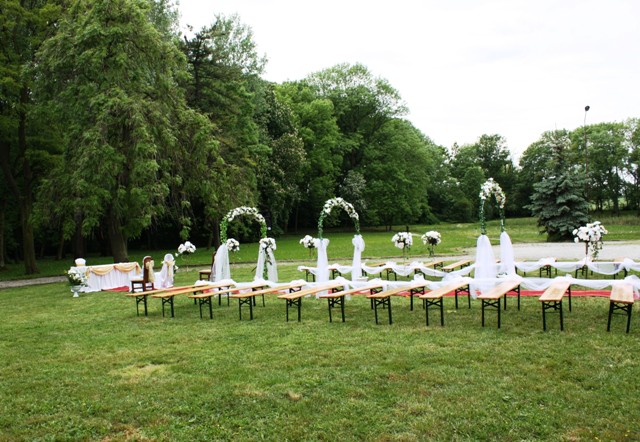 Uroczyste ustawienie rzędów drewnianych ławek i dekoracyjnych łuków z białymi kwiatami na trawniku, przygotowane na ceremonię ślubną w plenerze.