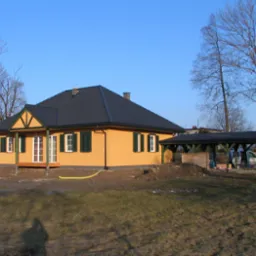 Żółty dom z ciemną dachówką i zielonymi okiennicami, z dobudowaną wiatą garażową. Widoczne drzewa bez liści i trawnik. W tle błękitne niebo.