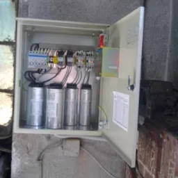 Otwarta szafa elektryczna z czterema srebrnymi kondensatorami cylindrycznymi, widoczne bezpieczniki, przekaźniki i okablowanie, zamontowana na szarej, chropowatej ścianie.