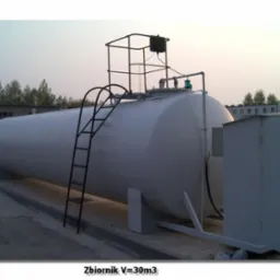Biały, stalowy zbiornik naziemny o pojemności 30m3 z drabinką i platformą inspekcyjną, umieszczony na betonowej podstawie przy budynku.