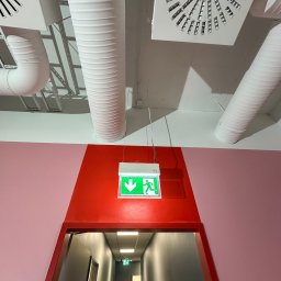 Paweł Rot - Czerwony portal z zielonym znakiem wyjścia awaryjnego, nad którym biegną białe, karbowane kanały wentylacyjne i kwadratowe nawiewniki sufitowe. Ściana obok jest różowa.