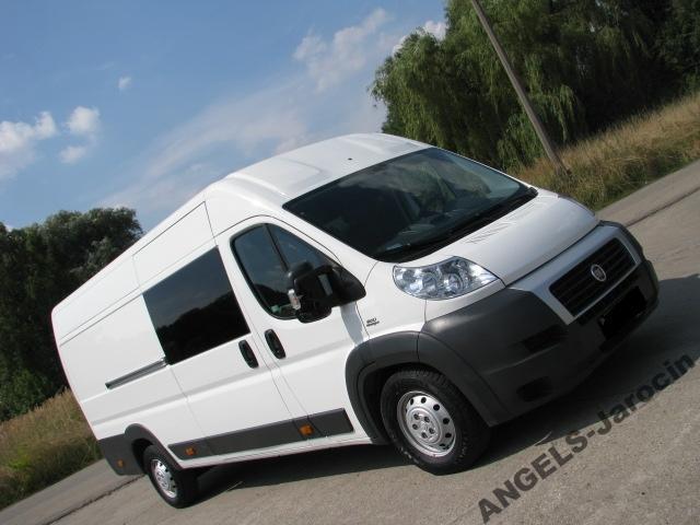 Biały samochód dostawczy Fiat Ducato z przyciemnianymi szybami, zaparkowany na asfaltowej drodze w słoneczny dzień.