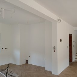 Kompleks pro group - Świeżo pomalowane na biało wnętrze z podłogą w jasną jodełkę, wystającymi przewodami elektrycznymi i częściowo wykończonymi drewnianymi schodami.