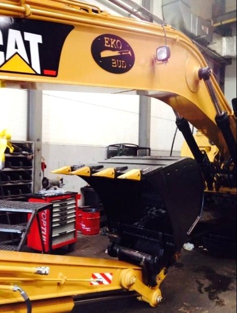 Żółta koparka marki CAT z czarnym logiem EKO BUD, zaparkowana w warsztacie z czerwonymi szafkami narzędziowymi w tle.