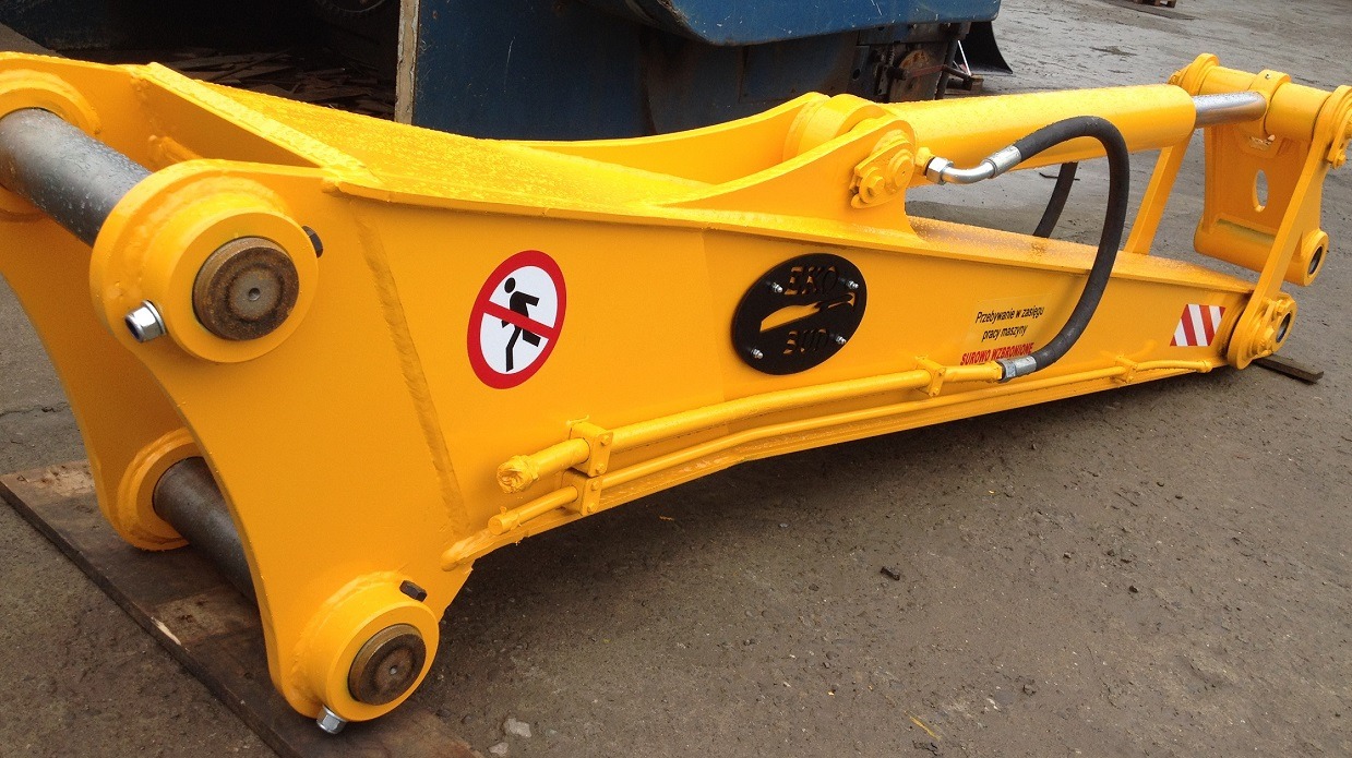 Żółty, hydrauliczny osprzęt do maszyny budowlanej, z widocznymi elementami mocującymi, w tym cylindrami hydraulicznymi, oraz naklejką ostrzegawczą z zakazem wchodzenia, na betonowym podłożu.