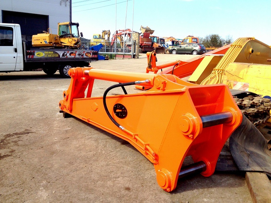 Pomarańczowe ramię koparki z hydraulicznym siłownikiem leżące na ziemi, w tle zaparkowane maszyny budowlane i samochód dostawczy z minikoparką na pace.