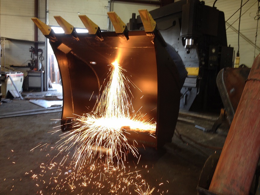 Spawanie łyżki koparki z widocznymi iskrami w warsztacie, żółte zęby łyżki, w tle maszyny przemysłowe.