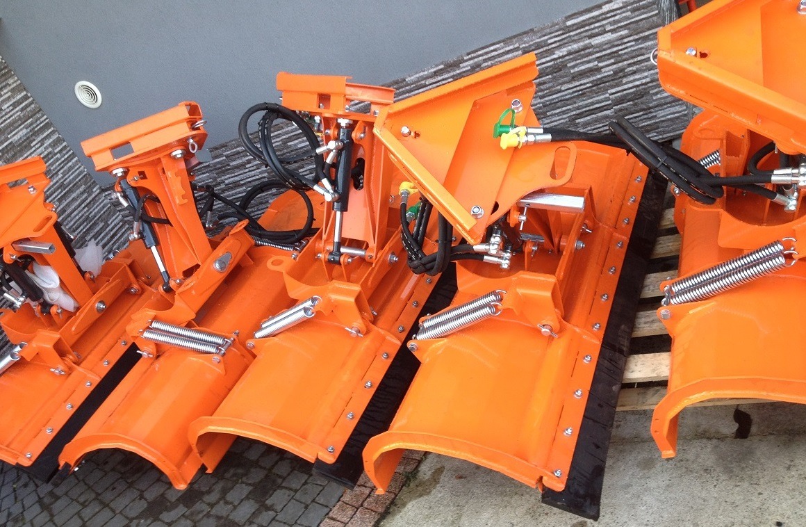 Rząd nowych, pomarańczowych pługów do odśnieżania z widocznymi sprężynami i przewodami hydraulicznymi, ustawionych na brukowanej powierzchni przed budynkiem.