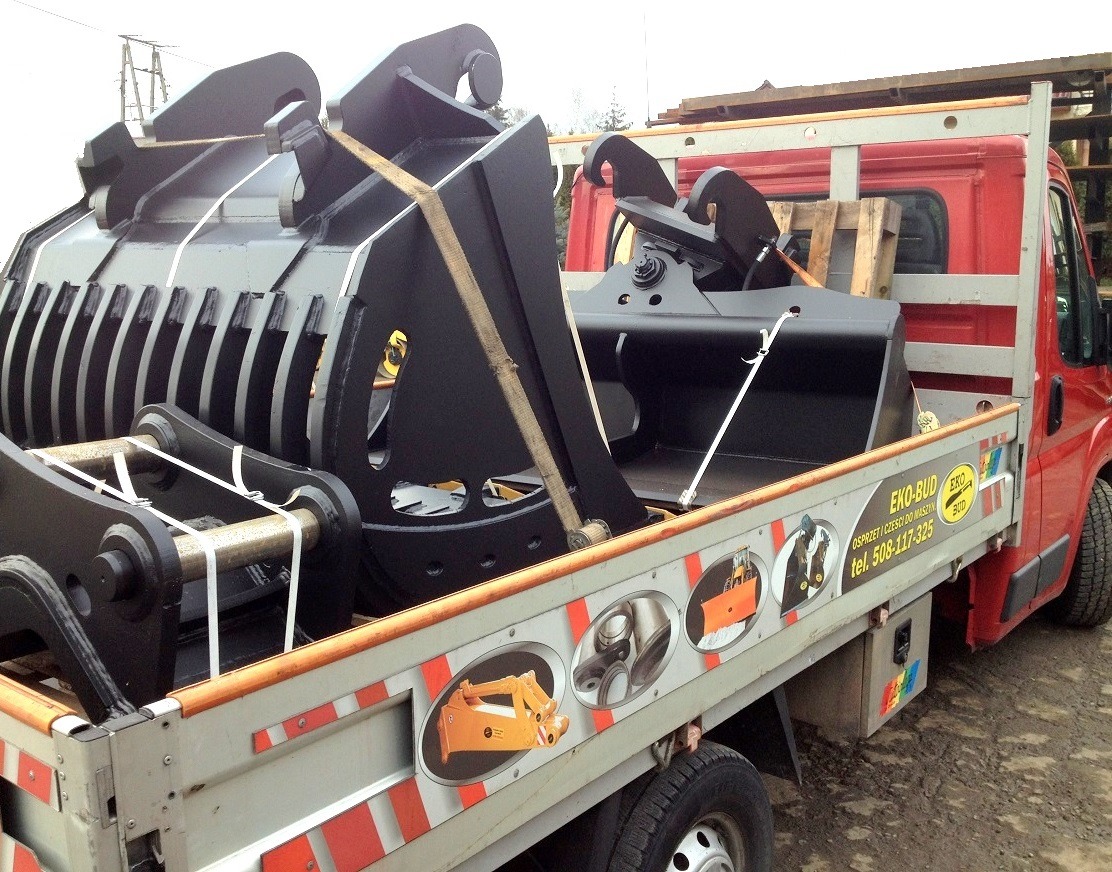 Czerwony samochód dostawczy z czarnymi łyżkami do maszyn budowlanych, zabezpieczonymi pasami transportowymi na pace. Widoczne logo firmy EKO-BUD na burcie.