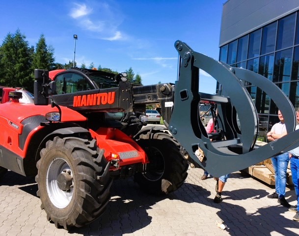 Czerwona ładowarka teleskopowa Manitou z osprzętem do drewna, widok z boku na tle budynku i drzew