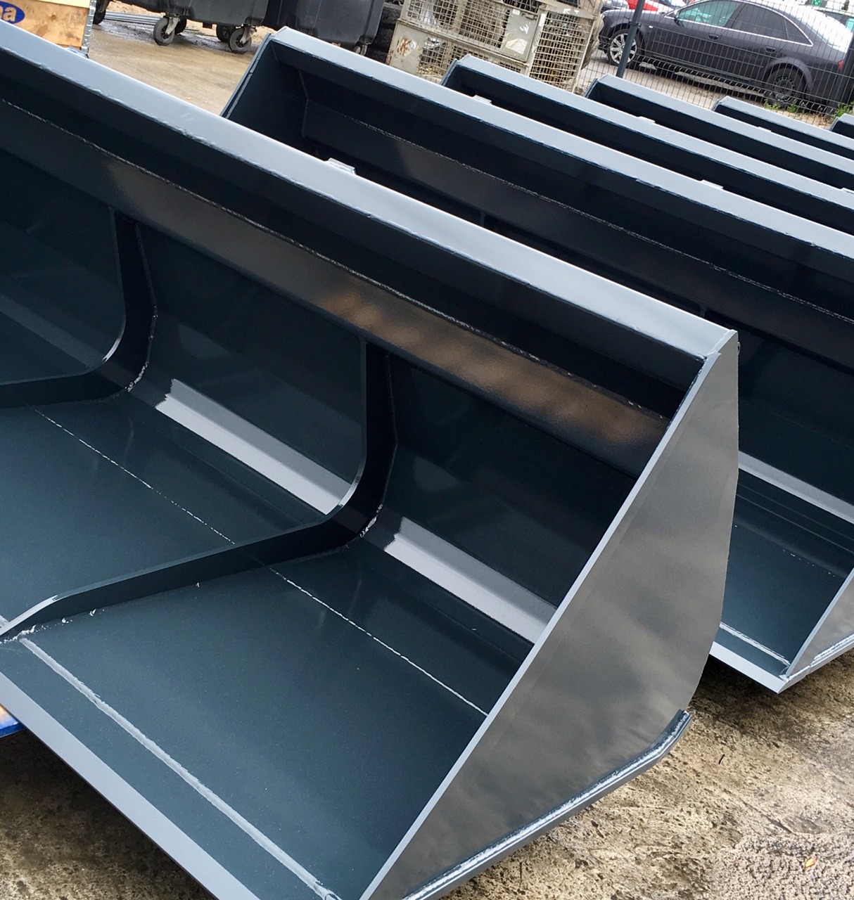 Nowe, pomalowane na szaro łyżki do koparek ustawione w rzędzie, widoczne ślady po spawaniu.