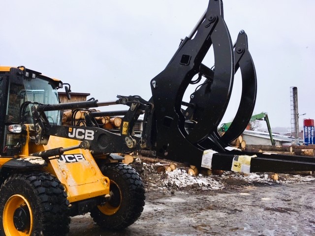 Żółta maszyna JCB z czarnym chwytakiem do drewna, w tle stosy pociętych pni drzew i budynki przemysłowe pod pochmurnym niebem.