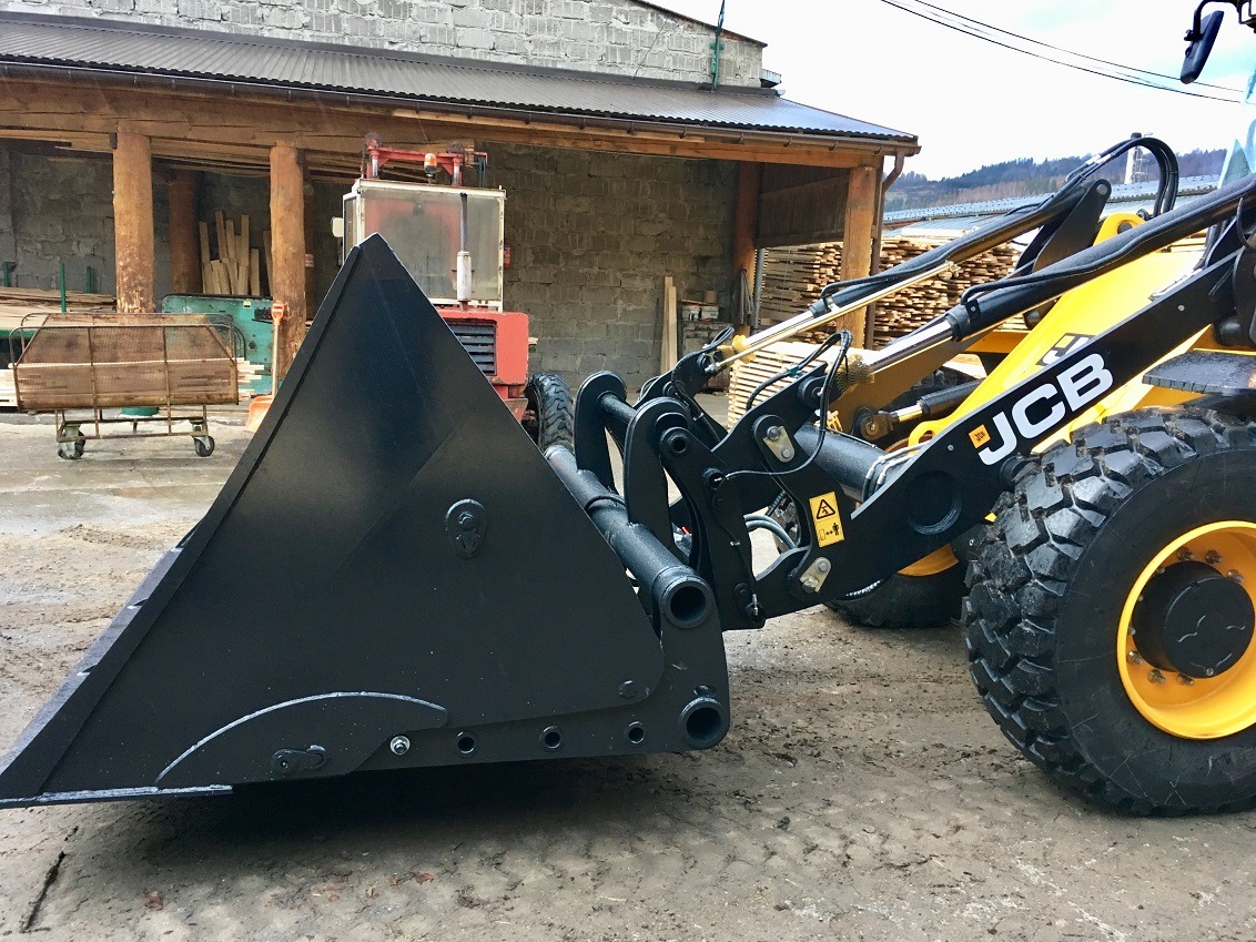 Czarny osprzęt ładowarki JCB z widocznym logo na tle budynku z drewnianymi elementami konstrukcyjnymi i wózkiem z drewnem.