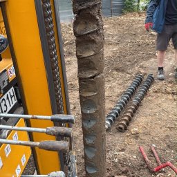 WM Geotechnika S.C. - Pionowy, zabłocony świder wiertniczy, częściowo wyciągnięty z ziemi przez żółtą maszynę z widocznymi dźwigniami sterującymi. Obok leżą zapasowe wiertła.