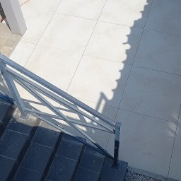 Katarzyna Hofman - Jasne płytki tarasowe w trakcie montażu, z pomarańczowym kablem i miotłą. Obok ciemne schody z metalową balustradą, pod konstrukcją rzucającą wzorzysty cień.