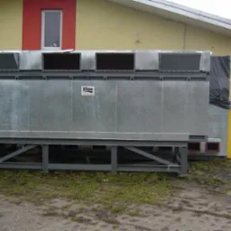 Duża, przemysłowa suszarnia biomasy z metalową obudową, widoczna na zewnątrz budynku z żółtą elewacją i czerwonym akcentem, z elementami wentylacyjnymi na górze i konstrukcją wsporczą na dole.