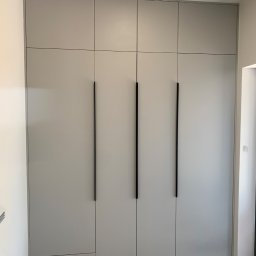 Ami Meble - Jasnoszara szafa wnękowa od podłogi do sufitu, z czterema pionowymi czarnymi uchwytami i modułowymi drzwiami, w minimalistycznym stylu.