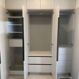 Ami Meble - Nowoczesna, jasnoszara zabudowa garderobiana z otwartymi drzwiami, prezentująca modułowe wnętrze z półkami, szufladami i drążkami. Widoczne czarne uchwyty i górne szafki.
