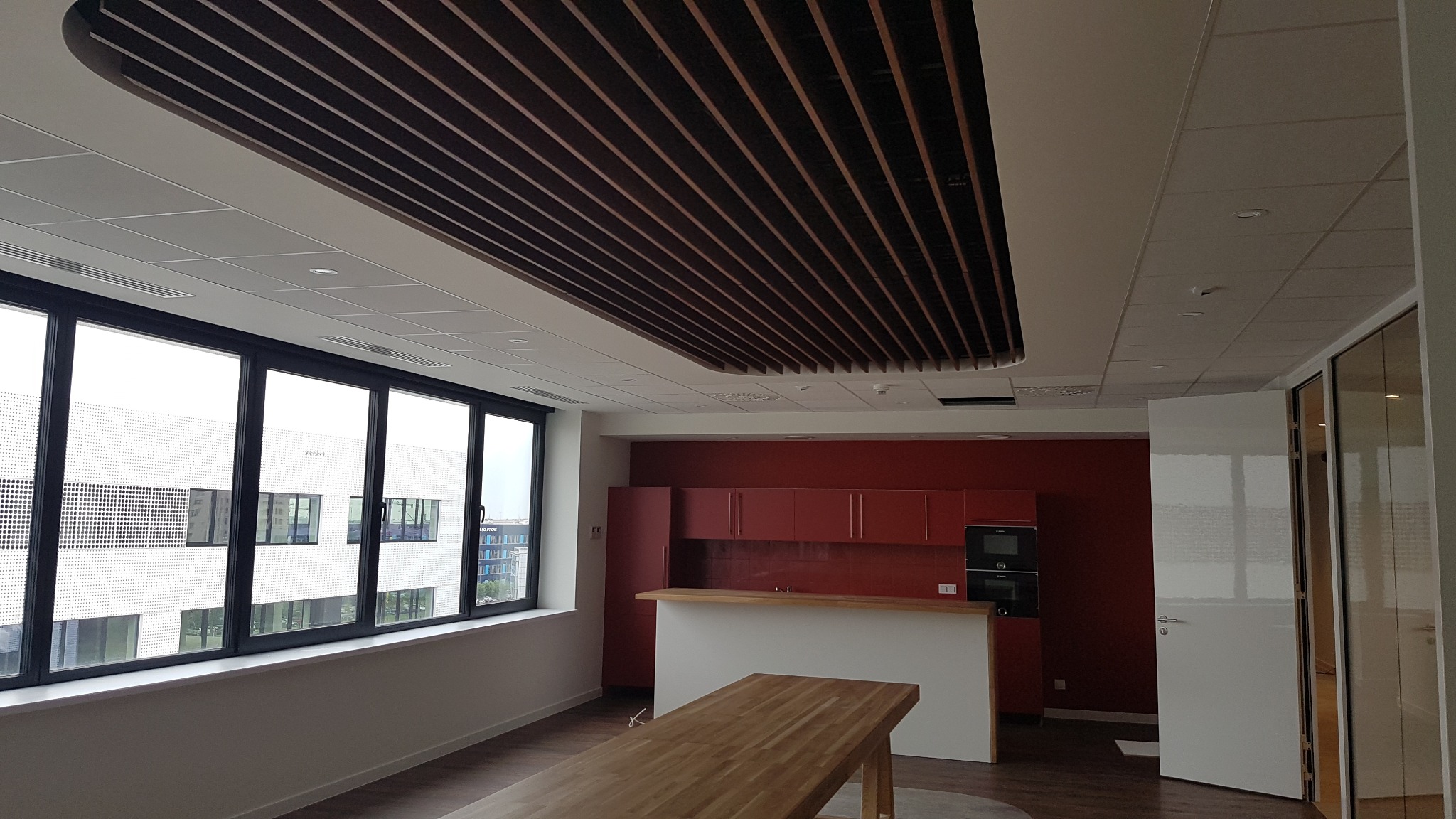Nowoczesne biuro z drewnianą podwieszaną konstrukcją sufitową, dużym oknem z widokiem na budynek biurowy, czerwonymi szafkami kuchennymi i drewnianym stołem.