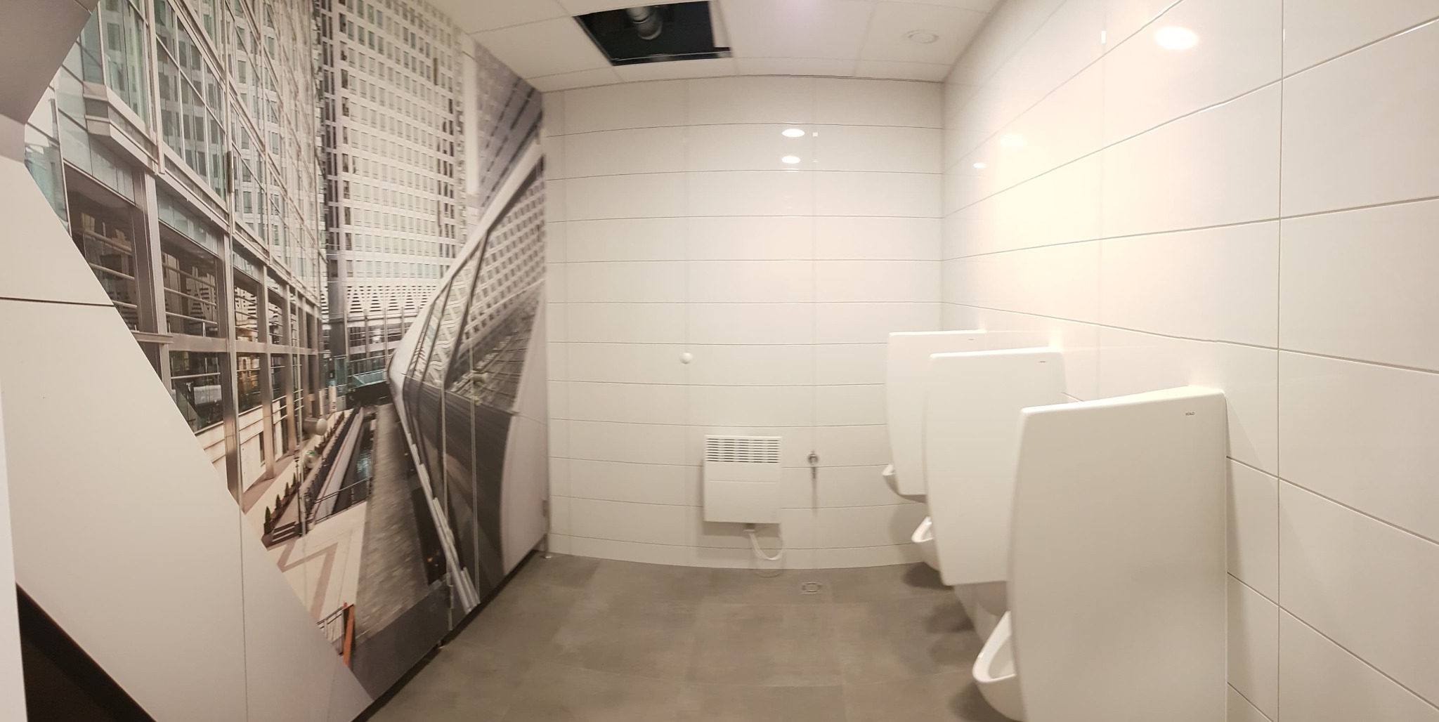 Publiczna toaleta męska z białymi pisuarami i fototapetą z nowoczesnym budynkiem biurowym na ścianie, grzejnik elektryczny na ścianie z białych płytek