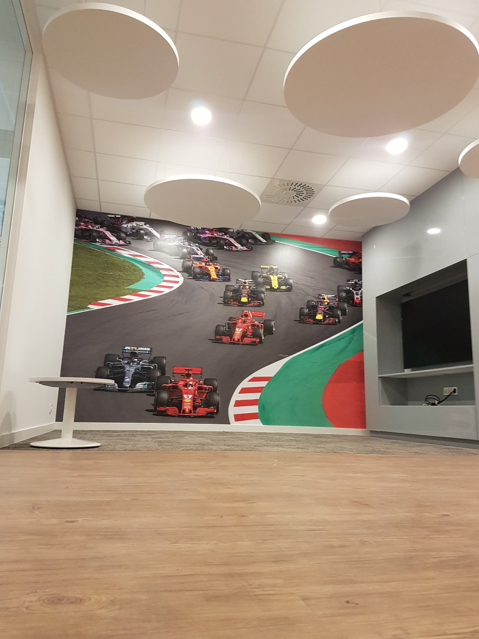 Wnętrze biura z fototapetą przedstawiającą wyścig Formuły 1, okrągłe panele akustyczne na suficie, jasna drewniana podłoga i minimalistyczny stolik.