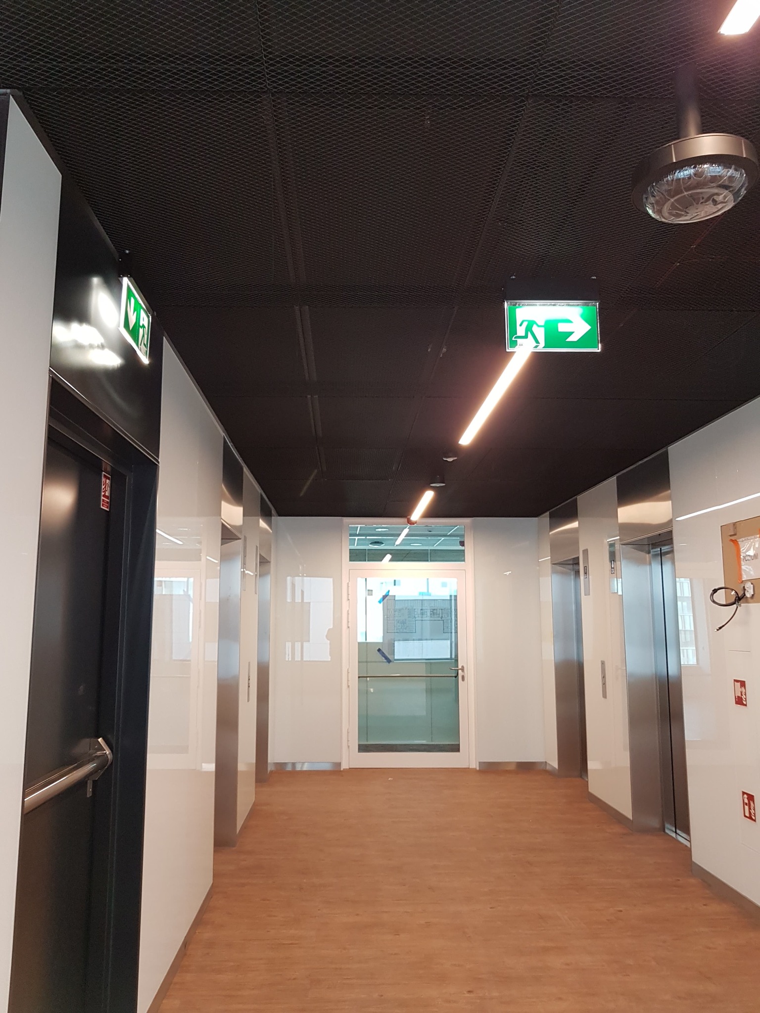 Korytarz biurowy w Krakowie z windami, oświetleniem LED i podwieszanym sufitem z siatki, widoczne znaki ewakuacyjne.