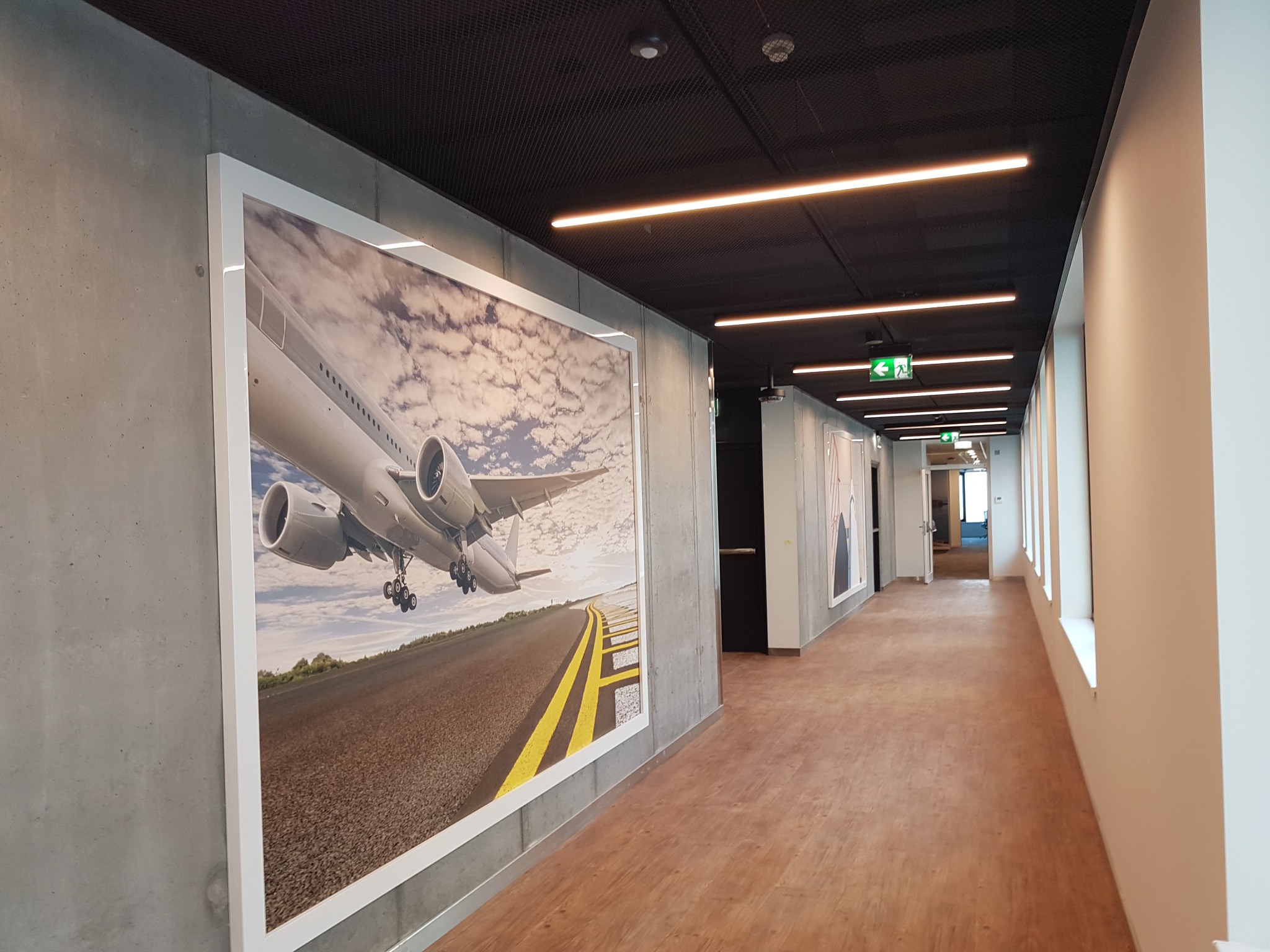 Długi korytarz biurowy z drewnopodobną podłogą, betonowymi ścianami i rzędem liniowych lamp sufitowych; na ścianie wisi duży oprawiony plakat z samolotem.