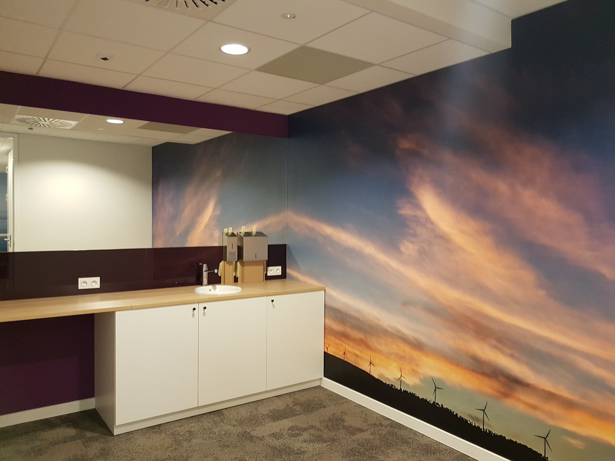 Nowoczesna łazienka biurowa z umywalką nablatową, białymi szafkami i blatem z jasnego drewna, uzupełniona fototapetą z motywem zachodu słońca i wiatraków na ścianie.