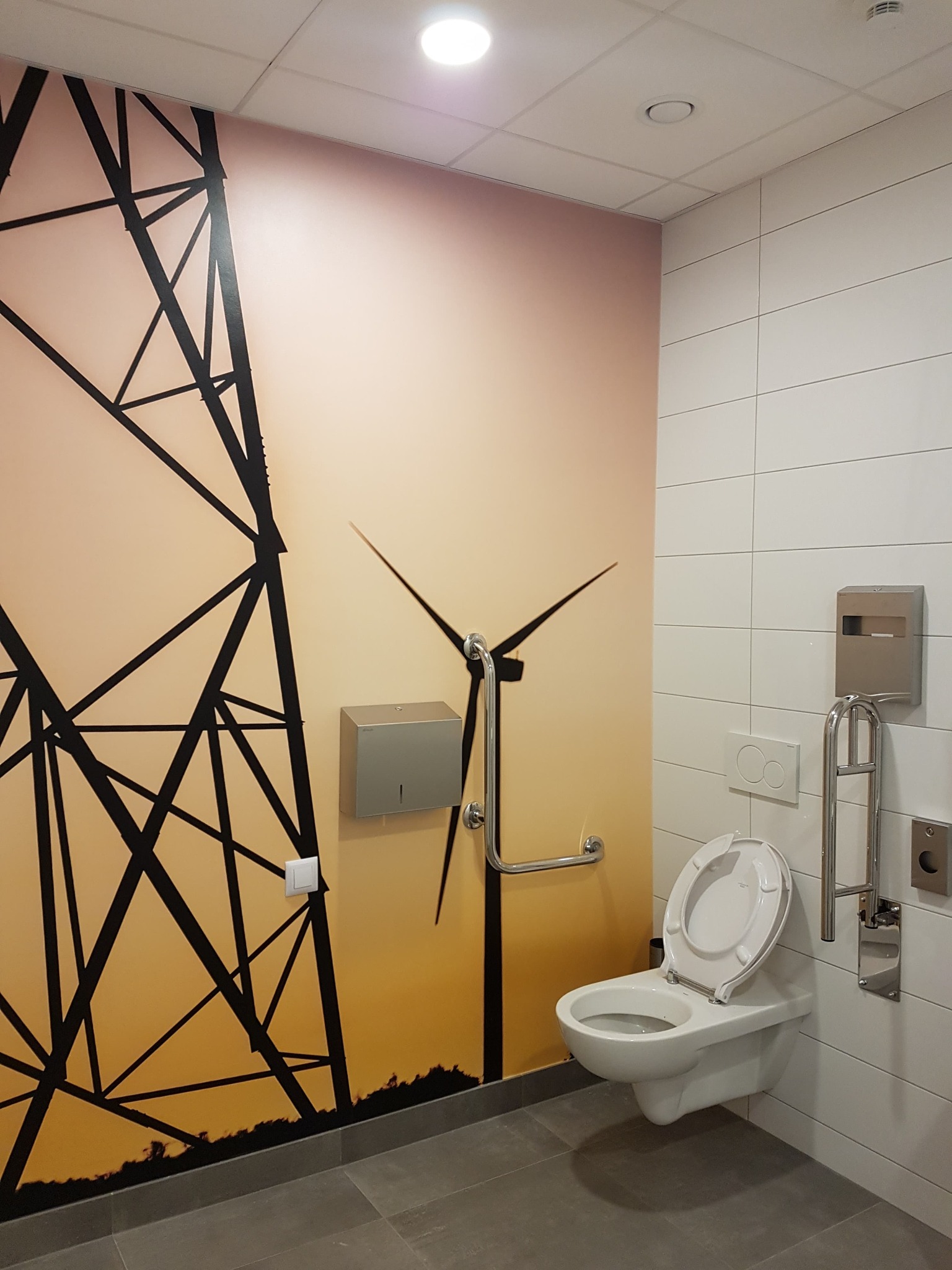 Toaleta w biurze z fototapetą przedstawiającą słup energetyczny i turbinę wiatrową, wyposażona w uchwyty dla niepełnosprawnych, białe płytki i szarą podłogę.