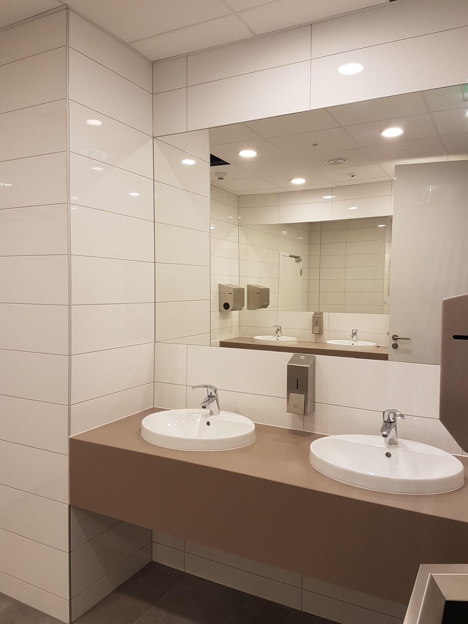 Publiczna toaleta z dwoma umywalkami, białymi płytkami na ścianach i dużym lustrem, w którym odbija się druga umywalka i dozowniki mydła.