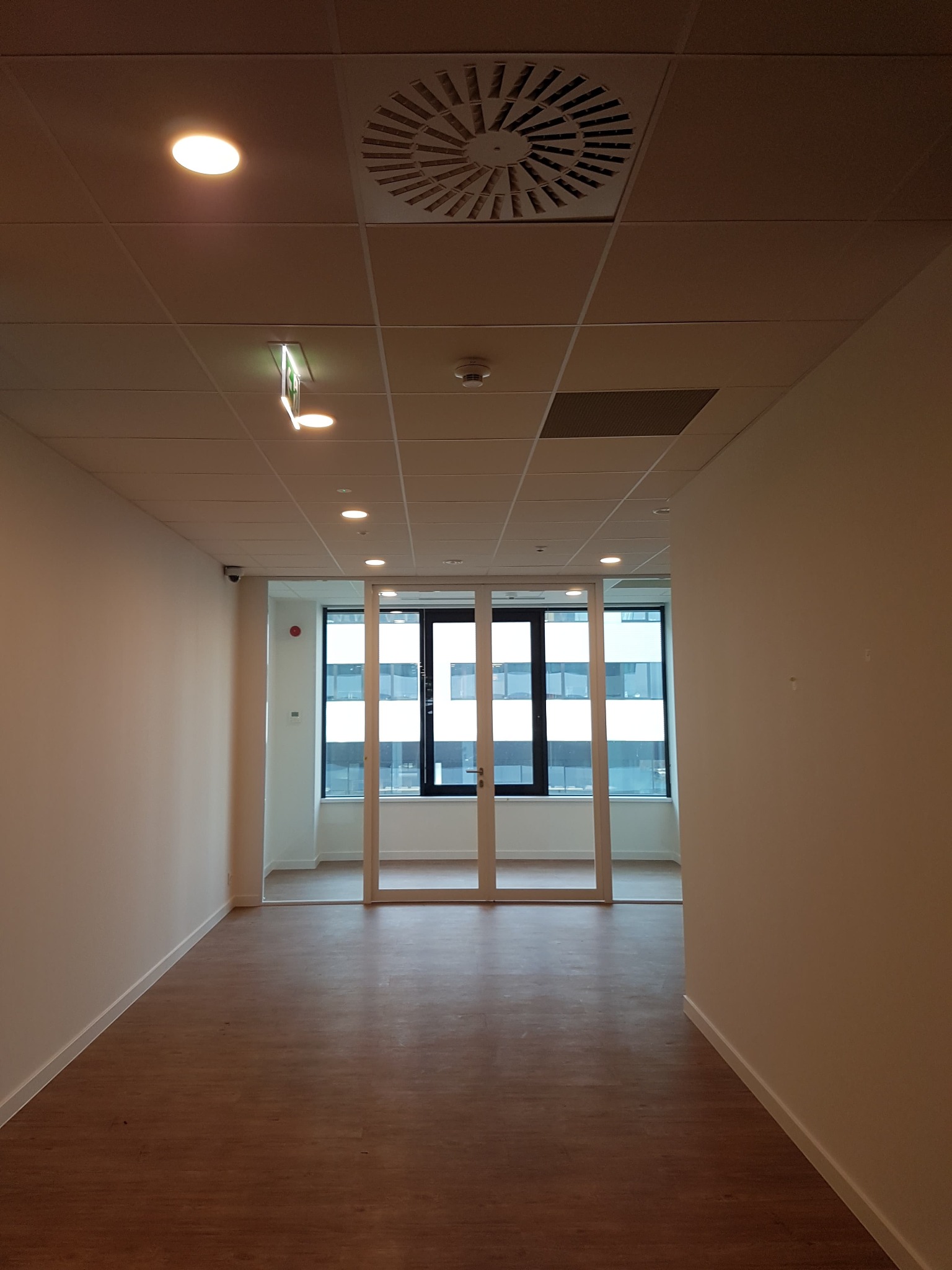 Wnętrze biura z panelami podłogowymi, ścianami pomalowanymi na jasny kolor i przeszklonymi drzwiami przesuwnymi w ramie, widok w głąb pomieszczenia z oknami.