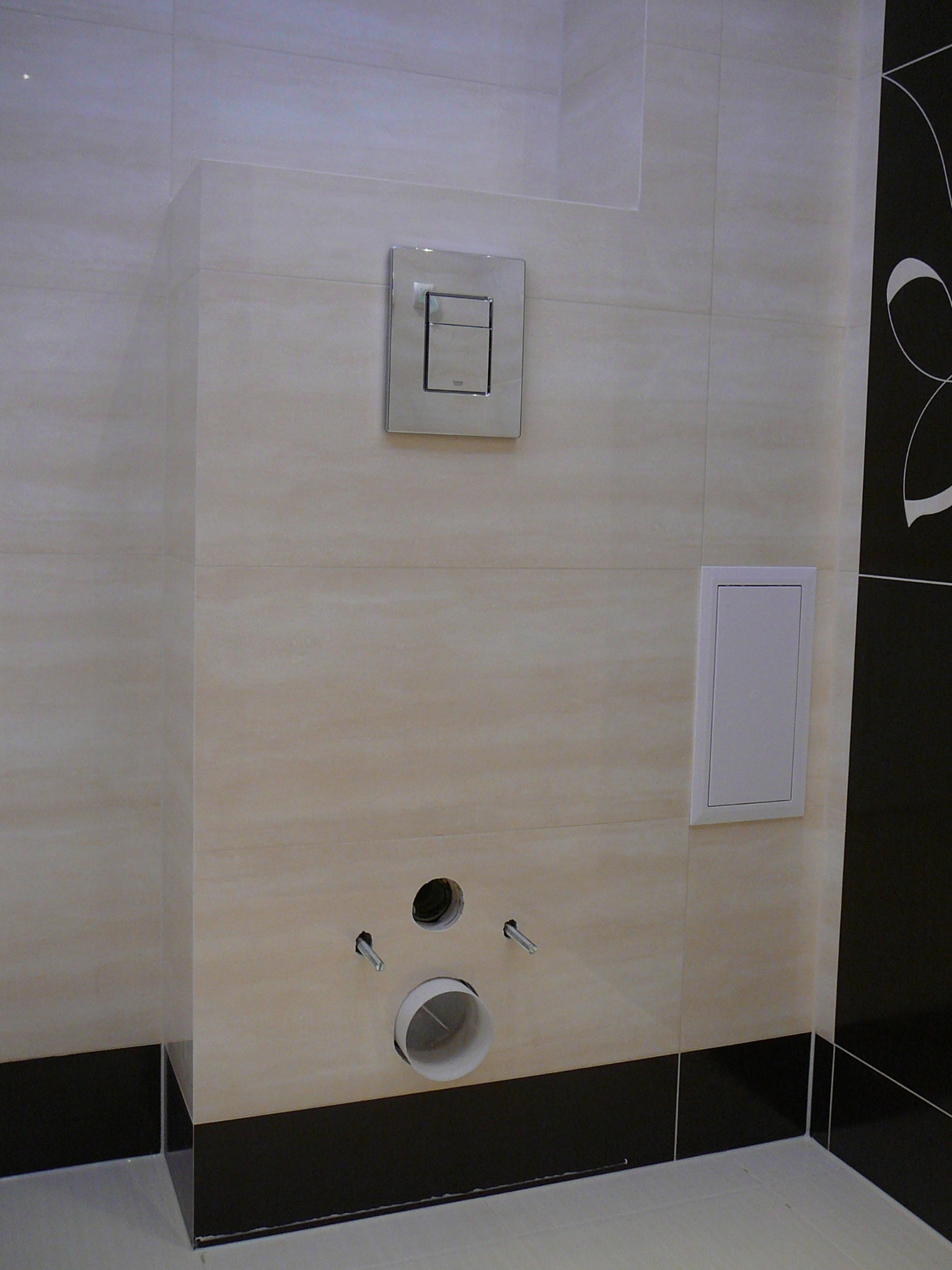 Ściana łazienki z beżowymi płytkami, widoczne otwory na instalację toalety podtynkowej, przycisk spłukiwania i rewizja, czarny pas płytek u dołu.