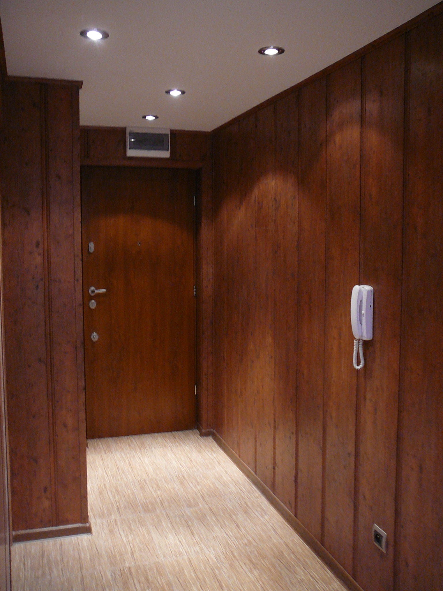 Korytarz z panelami ściennymi w odcieniach brązu, jasną podłogą imitującą drewno i białą słuchawką domofonu na ścianie.