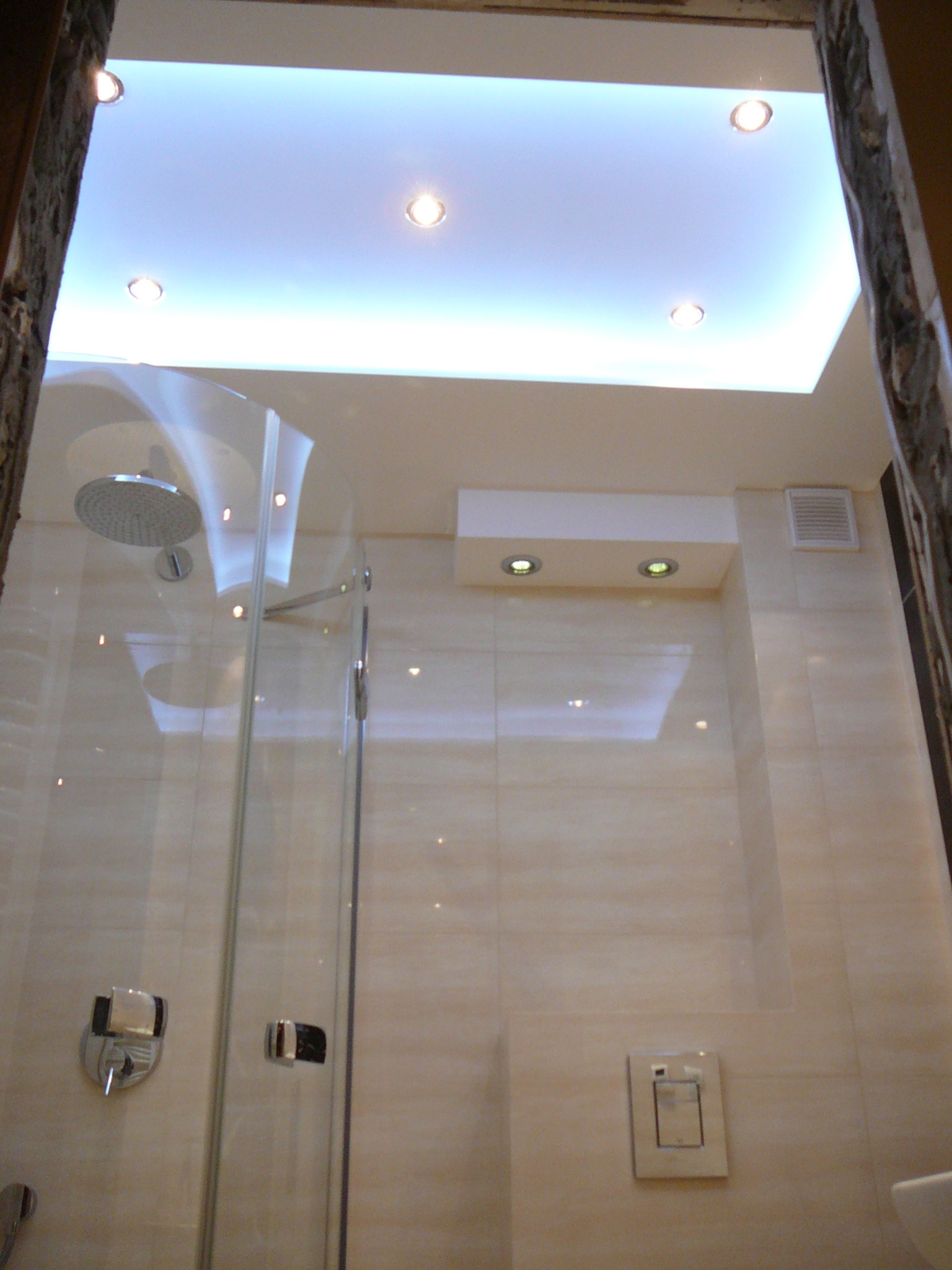 Nowoczesna kabina prysznicowa ze szklanymi drzwiami i deszczownicą w jasnej łazience z podwieszanym sufitem i oświetleniem punktowym.