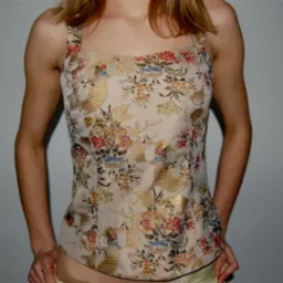 Kobieta prezentuje top z kwiecistego, brokatowego materiału z grubymi ramiączkami, na tle szarej ściany z widoczną dekoracją.