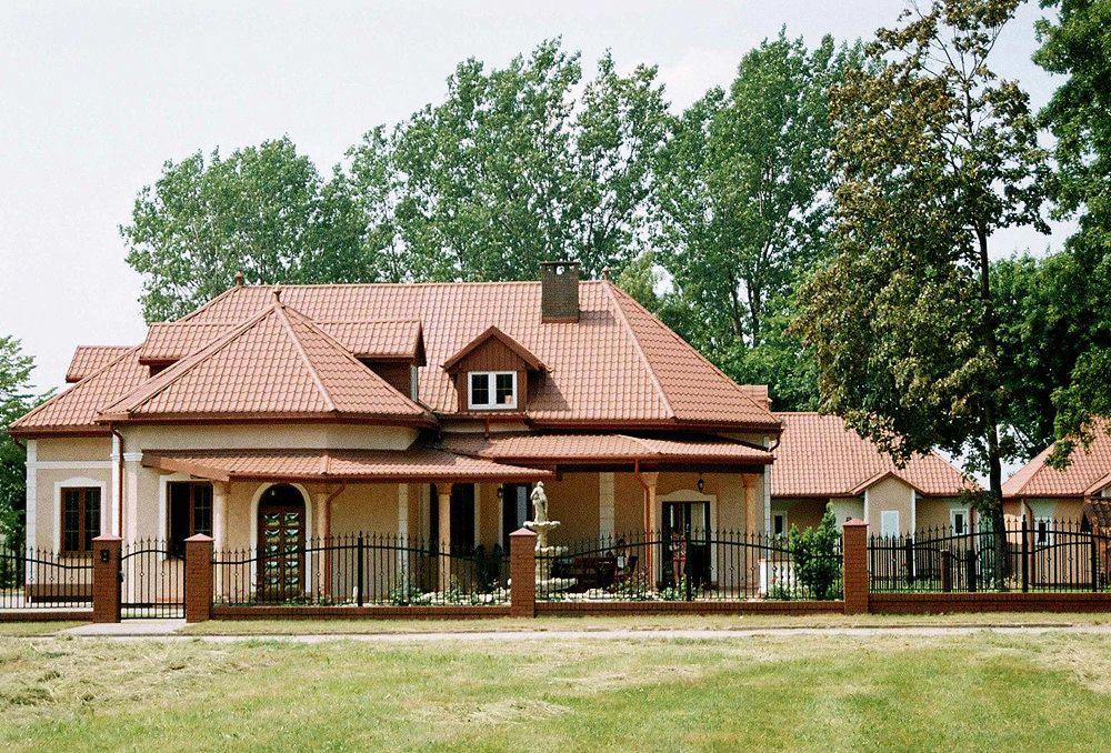 Rozległa rezydencja z czerwoną dachówką, otoczona czarnym, kutym ogrodzeniem i zielonym trawnikiem, z widoczną fontanną przed wejściem.