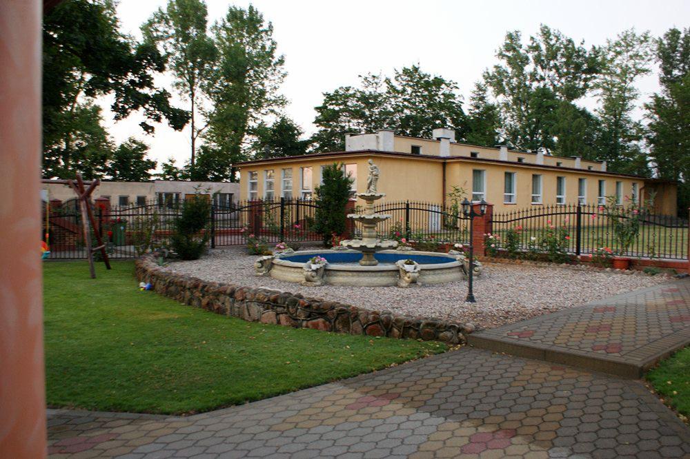 Dwukondygnacyjny budynek z beżową elewacją, otoczony czarnym, kutym ogrodzeniem, z fontanną z posągiem pośrodku dziedzińca, widok z boku na trawnik, kamienny murek i brukowany chodnik.